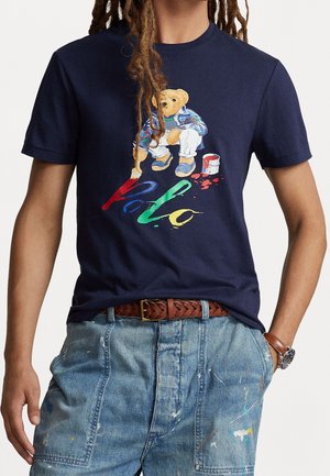 Marineblå t-shirt med en bjørnemaleri og flerfarvet "Polo" tekst, båret med malersprøjtet denimshorts, flettet bælte og ur.
