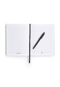 Bolígrafo negro colocado sobre un cuaderno abierto con páginas rayadas. El cuaderno tiene un marcador de cinta negra y contiene una pequeña ilustración de un velero.