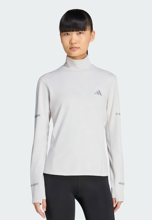 Ung kvinde med glat sort hår iført en hvid langærmet rullekrave og sorte leggings, stående mod en ensfarvet baggrund.