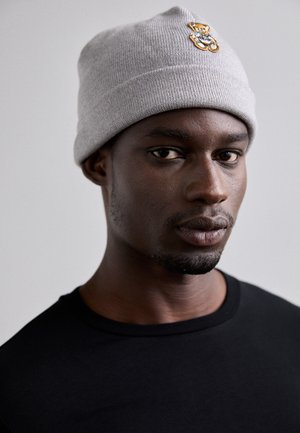 Bonnet en tricot gris avec un patch en or en forme d'ours, ajusté près de la tête. Porté avec un t-shirt noir à col rond. Texture lisse, design décontracté.
