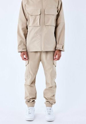 Veste zippée beige avec deux poches avant, associée à un pantalon fuselé assorti avec des poches cargo et des poignets élastiques. Baskets blanches visibles.