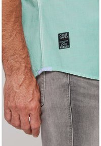 Camicia verde menta a maniche corte con cuciture bianche e un'etichetta in tessuto nera. Pantaloni in denim grigio parzialmente visibili. Mano appoggiata sulla coscia.
