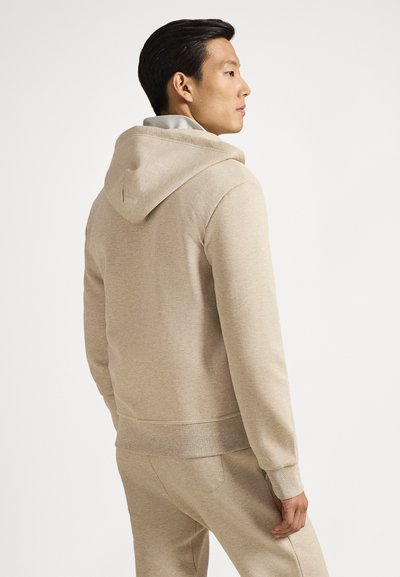 Μπεζ φούτερ με κουκούλα, μαλακή υφή, εφαρμοστά μανσέτες και τσέπη καγκουρό, εμφανίζεται σε ένα άτομο από ελαφριά πλάγια όψη.