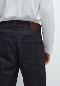 Jeans in denim scuro con taglio dritto, dotati di due tasche posteriores e un'etichetta in pelle marrone alla vita. Dettaglio di cucitura semplice.