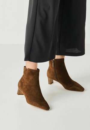 Brown suede ankle boots with block heels and side zippers worn with wide black cropped pants on a white floor.