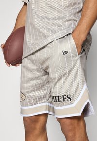 Beige mesh shorts with white stripes, elastic waistband, side pockets, and “CHIEFS” in black lettering. A football is held in the left hand.