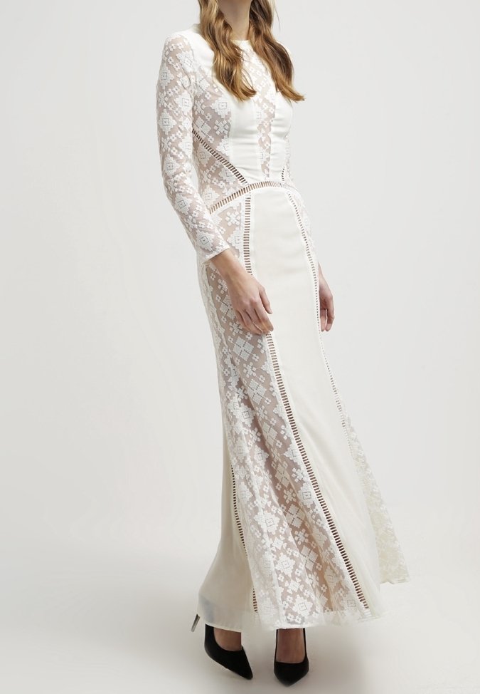 Femme portant une longue robe en dentelle blanche avec des motifs géométriques et des panneaux, associée à des escarpins noirs à bout pointu, sur un fond uni.