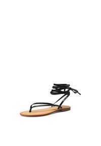 Sandali neri piatti con un design a striscia minimale e una suola beige. Le cinghie sono flessibili e si avvolgono attorno alla caviglia per una vestibilità sicura.