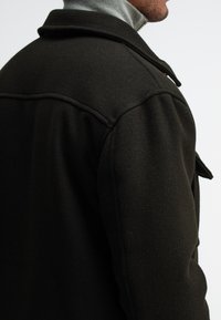Dark wool coat with a textured finish, featuring a folded collar and shoulder seams, shown from the right side.