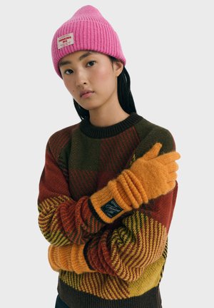 Jeune femme portant un bonnet en tricot rose, un pull à carreaux multicolore et des gants en tricot orange, les bras croisés devant un fond uni.