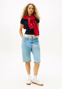 Bermuda shorts in denim blu con una vestibilità comoda, abbinati a una t-shirt attillata navy e a una felpa rossa drapesata sulle spalle. Sneakers bianche.