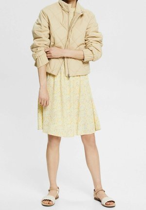 Donna con giacca trapuntata beige, abito giallo a fiori fino al ginocchio e sandali bianchi con cinturino intrecciato, in piedi con le braccia incrociate.