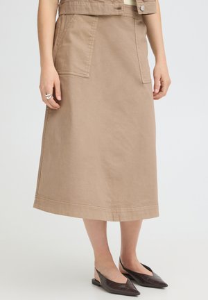 Femme portant une jupe midi beige avec des poches devant, associée à des ballerines slingback pointues marron foncé, mains détendues sur les côtés.