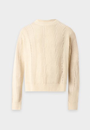 Homme portant des lunettes de soleil noires, un pull en tricot crème, un jean bleu clair et des chaussures noires, debout sur un fond clair uni.