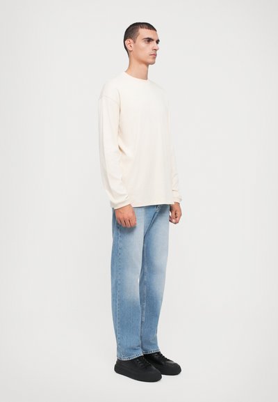 Beige long-sleeve t-shirt paired with light blue jeans. The outfit features a relaxed fit and black low-top sneakers. Simple design.