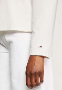 Camicia a maniche lunghe in cotone color crema con una consistenza morbida, caratterizzata da un piccolo ricamo a forma di bandiera sul polsino, abbinata a pantaloni bianchi.