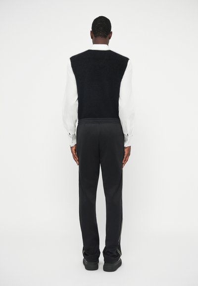 Čierny bezrukávový sveter cez bielu dlhú tričko s dlhým rukávom, doplnený o čierne športové nohavice so bočnými pruhmi. Hladké textúry a voľný strih.