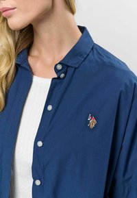 Tumedat sinine, nööbidest särgiga särk, millel on krae, hõbedased nööbid ja väike tikand logo vasakul rinnal.