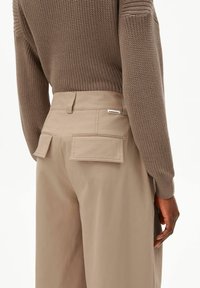 Persoon draagt beige hooggetailleerde broek met klepzakken en een taupekleurige gebreide trui, hand langs het lichaam rustend.