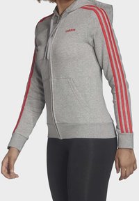 Felpa grigia con zip e cappuccio, caratterizzata da design a tre strisce rosse sulle maniche, cappuccio con cordini e tasche frontali. Realizzata in un morbido tessuto testurizzato.