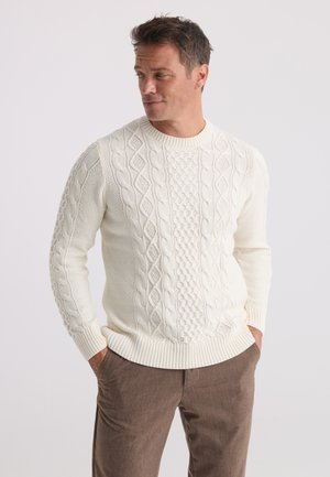 Homme portant un pull en maille torsadée blanc et un pantalon marron, les mains dans les poches, regardant sur le côté sur un fond uni.