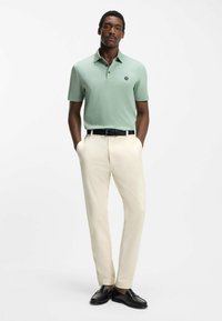 Mand iført lysegrøn kortærmet polo, beige bukser, sort bælte og sorte loafers, stående med hænderne i lommerne på hvid baggrund.