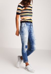 Suéter de manga corta a rayas en rosa, verde y marino, combinado con jeans rasgados de un azul claro con parches y zapatillas blancas.