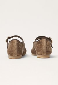 Vista posterior de zapatos de ante marrón con forma redondeada, una correa elástica flexible y un pequeño acento metálico circular. Suelas de goma planas.