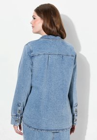 Camisa de mezclilla azul clara de manga larga, con puños abotonados y un corte relajado. Presenta un yugo trasero y detalles de costura.