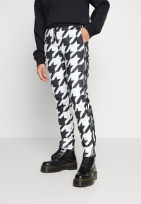 Kalhoty s černobílým kostkovaným vzorem ve stylu houndstooth, mají přiléhavý střih, šňůrkový pas a boční pruhy, doplněné černými šněrovacími botami.