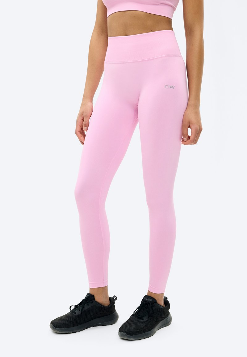 Γυναίκα που φοράει ροζ κολάν με ψηλή μέση και ασορτί αθλητικό σουτιέν, συνδυασμένα με μαύρα αθλητικά παπούτσια, στέκεται μπροστά σε λευκό φόντο.