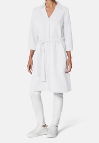 A person wears a white button-up dress with a tie belt, paired with white pants and sneakers, set against a plain background.