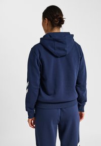 Frau mit dunklem Haar im Dutt, die einen navyblauen Hoodie und passende Hose trägt, von hinten vor einem schlichten weißen Hintergrund gesehen.