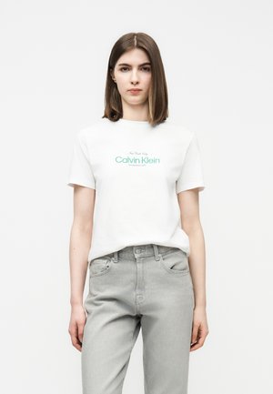 Νεαρή γυναίκα που φοράει λευκό T-shirt Calvin Klein με πράσινο λογότυπο και ανοιχτό γκρι ψηλόμεσο τζιν, στέκεται μπροστά σε ένα απλό λευκό φόντο.