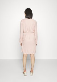 Vestido rosa, texturizado, com mangas longas e cintura com cordão, cai até meio da coxa. Usado com sapatos de salto bloco em creme. Fundo branco.