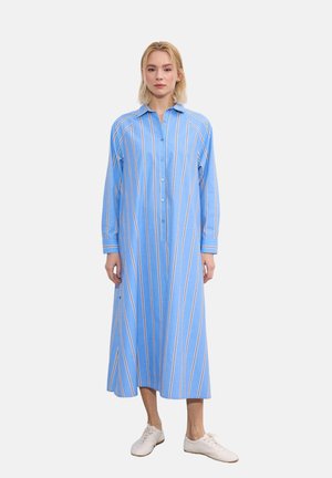 Žena nosi dlhé modré šaty s bielymi a béžovými vertikálnymi pásmi, zapínané vpredu na gombíky, s dlhými rukávmi a bielou ležérnou obuvou, stojaca.