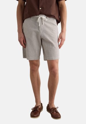 Hombre con pantalones cortos beige con rayas blancas y cordón, camisa marrón de manga corta y zapatos náuticos de cuero marrón, de pie sobre un fondo blanco.