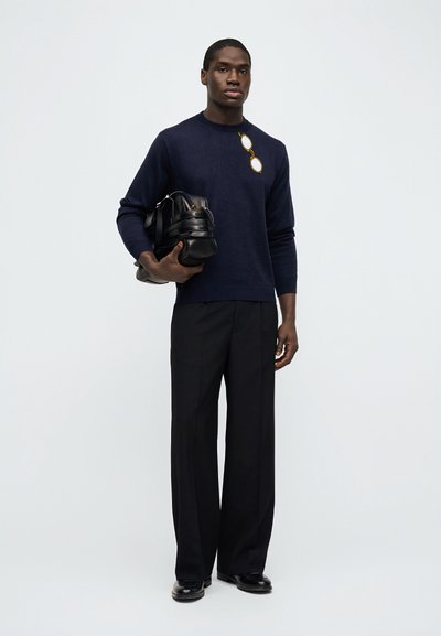 Άνδρας που φοράει μπλε ναυτικό πουλόβερ με κεντημένα γυαλιά, μαύρο παντελόνι φαρδύ, μαύρα παπούτσια, κρατώντας μια μαύρη δερμάτινη τσάντα duffle.