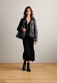 Svart läderbikersjacka över en svart stickad klänning, parat med ankelstövlar och en strukturerad handväska, presenterad mot en ljusgrå bakgrund.