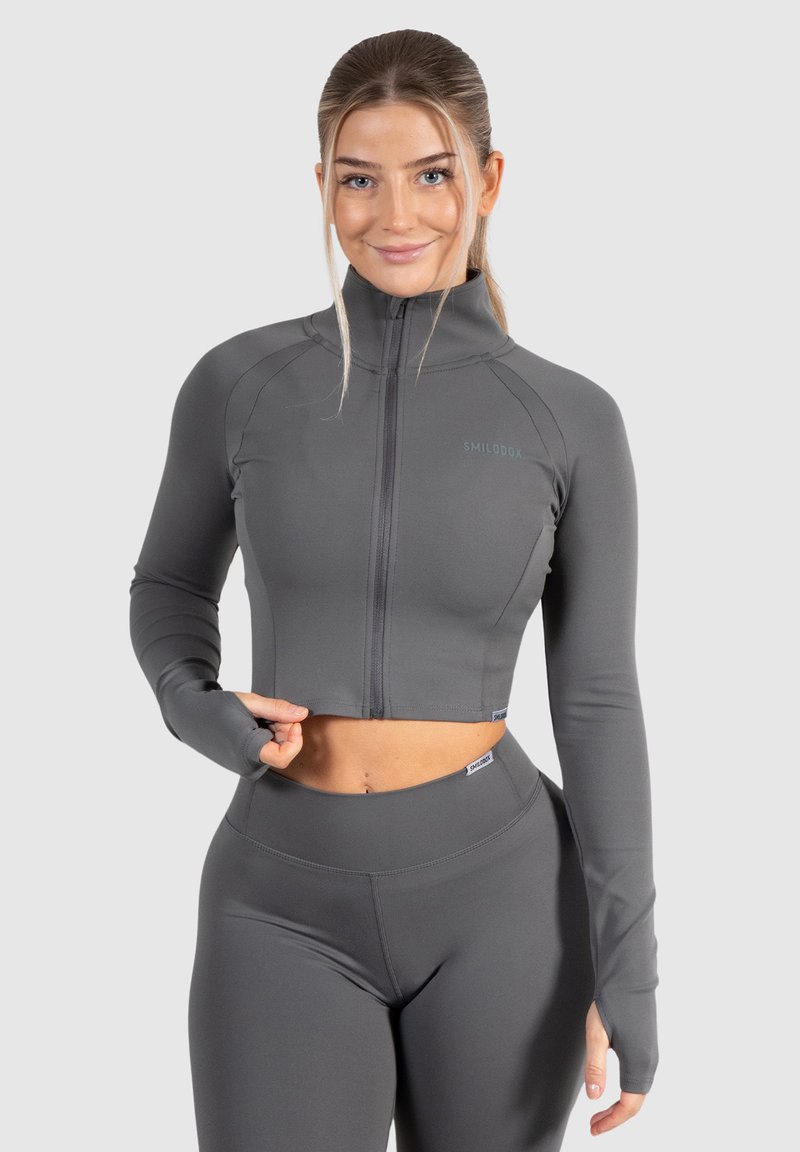 Graue, verkürzte Sportjacke mit langen Ärmeln, Halbreißverschluss vorne und einem glatten, dehnbaren Material. Merkmale ein Logo auf der Brust und Daumenlöcher.