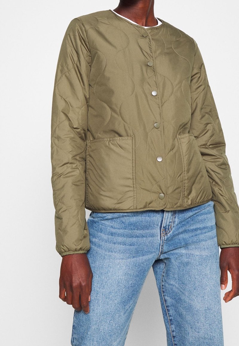 Personne portant une veste matelassée boutonnée vert olive avec deux poches avant et un jean en denim bleu clair sur un fond clair uni.
