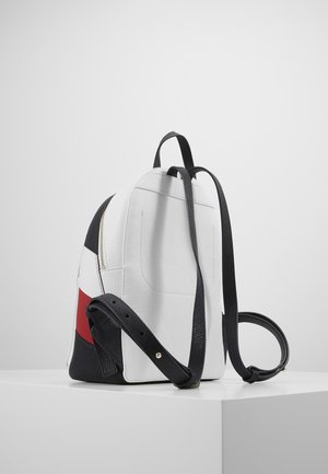 Kleiner weißer Rucksack mit schwarzen verstellbaren Gurten und einem Seitenpanel mit schwarzen, weißen und roten Farbblöcken, auf einer weißen Oberfläche platziert.