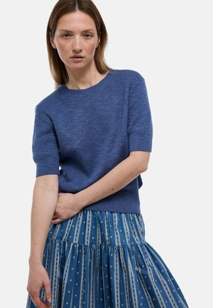 Femme aux cheveux bruns raides portant un haut en maille bleu à manches courtes et une jupe plissée bleue à motifs, debout devant un fond uni.