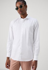 White long-sleeve button-up shirt with a classic collar. Smooth fabric, fitted shape, paired with light pink pants and round black sunglasses.