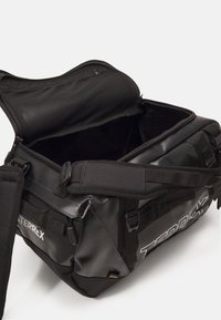 Le sac de sport noir présente un extérieur texturé, un compartiment supérieur zippé et une sangle ajustable. À l'intérieur, il est spacieux avec une poche en maille pour l'organisation.