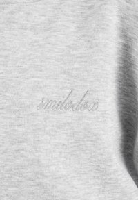 Jasnoszary materiał z subtelnym, haftowanym kursywą napisem „smilecloxi” wtapiającym się w tkaninę w okolicy klatki piersiowej.