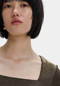 Primer plano de una persona que lleva una blusa de escote cuadrado en tono dorado metálico. Piel suave con un brillo sutil y cabello oscuro, acentuado por pendientes dorados.