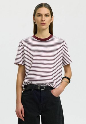 Femme aux cheveux bruns raides portant un t-shirt rayé blanc et bordeaux rentré dans un pantalon noir avec ceinture noire, debout devant un fond uni.