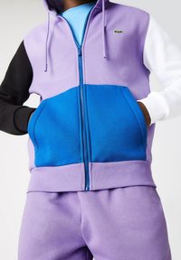 Felpa con zip a blocchi di colore, realizzata in tessuto morbido; presenta pannelli viola, neri e blu, con tasca a marsupio e cappuccio con cordino.