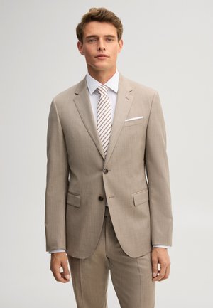 Man wearing a beige suit with a white shirt and striped tie standing against a plain light background.
